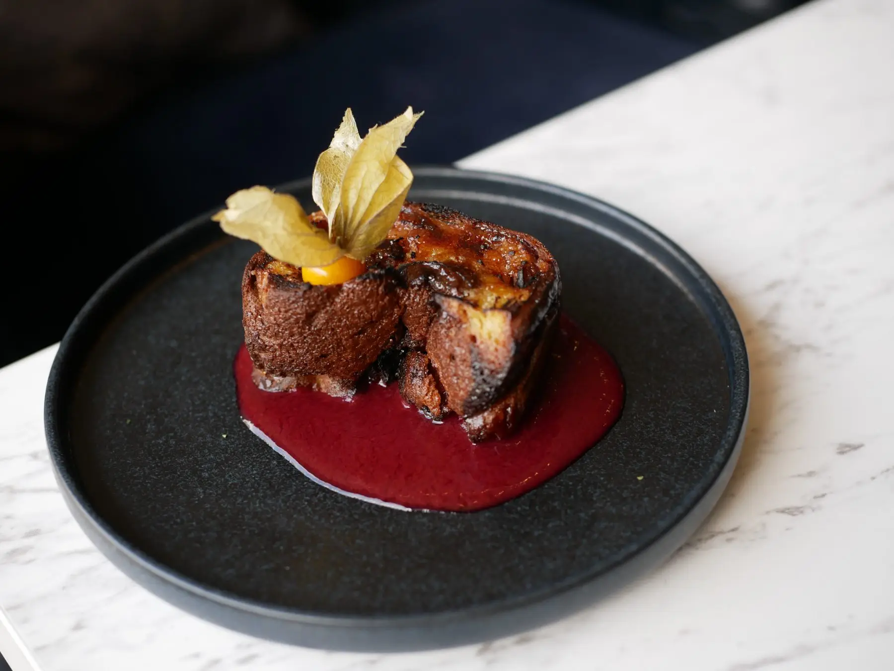 Dessert du Café Gourmand : brioche caramélisée sur un coulis de fruits rouges, assiette noire.