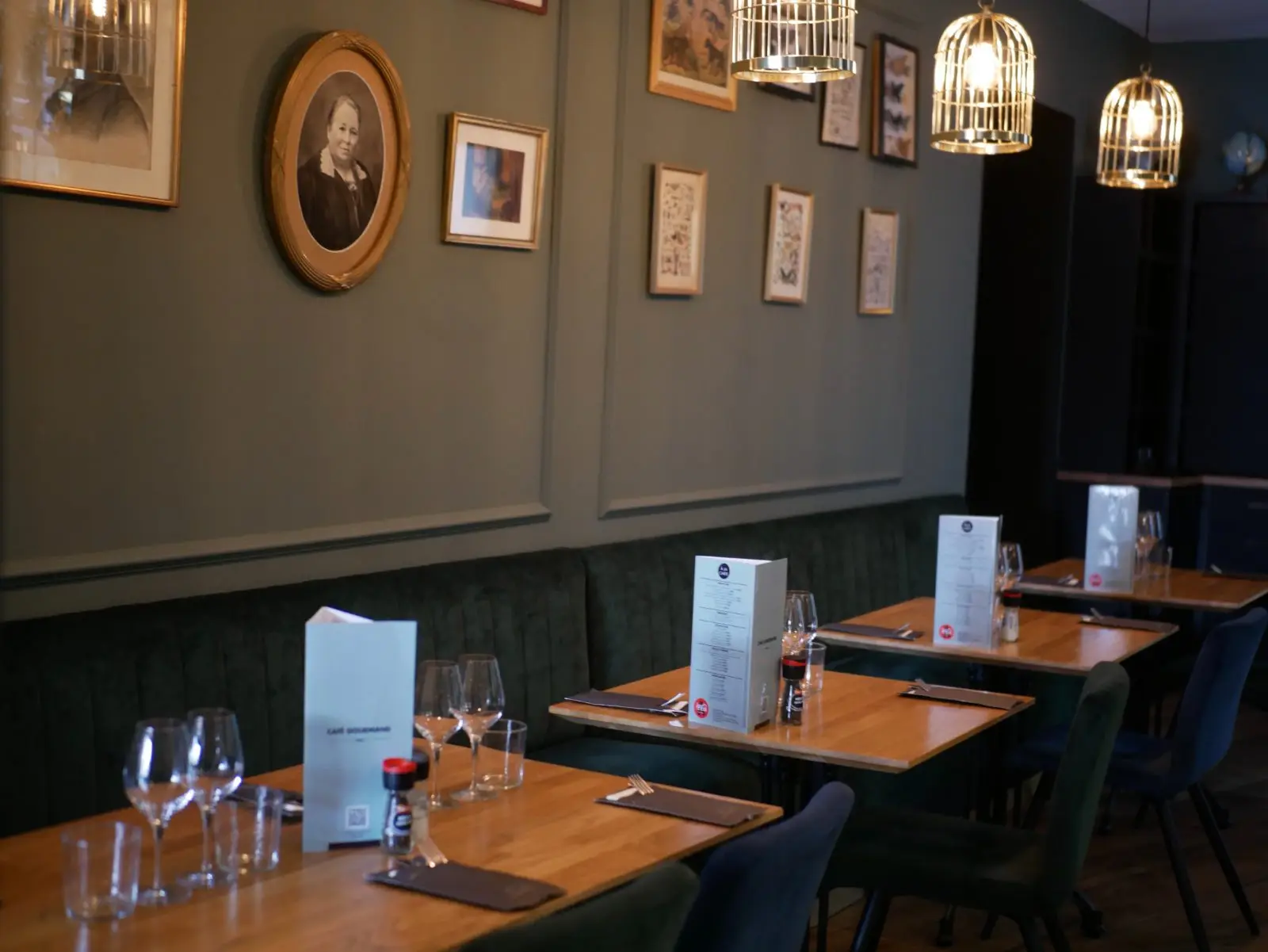 Salle du Café Gourmand : banquette de velours vert, tables dressées et cadres anciens sous des suspensions en laiton.