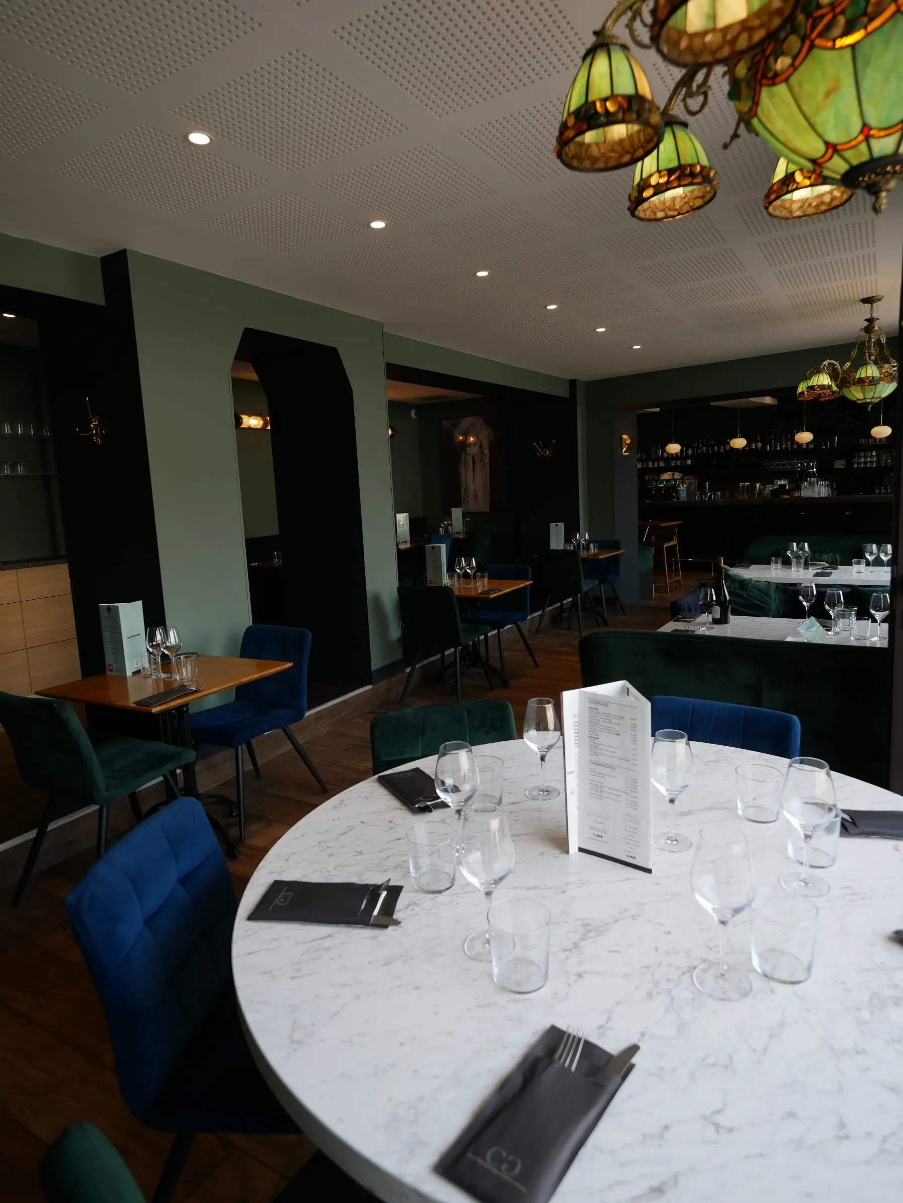 Salle du Café Gourmand : tables de marbre dressées, fauteuils de velours vert et bleu, suspensions Tiffany et bar en fond.